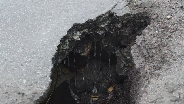Close-up pit on the asphalt caused by bad weather, traffic and poor maintenance. Water flows into a hole on the asphalt and drips down during the rain. A hollow in the ground in the middle of the road