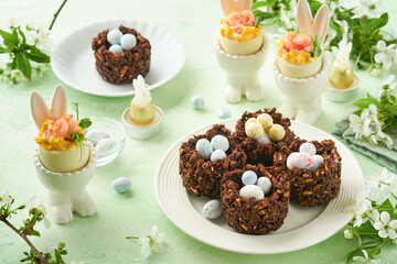 Easter chocolate nest cake with mini chocolate candy eggs with blossoming cherry or apple flowers on green background table. Creative recipe for Easter table with holiday decorations. Top view.