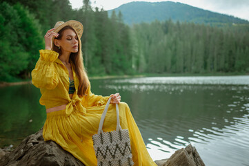 Fashionable woman wearing wicker hat, summer yellow suit with blouse and long skirt, carrying stylish bag, posing near beautiful lake in mountains. Copy, empty space for text