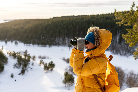 Young woman in yellow looking through binoculars at birds on snowy river against winter forest Birdwatching, zoology, ecology Research in nature, observation of animals Ornithology copy space