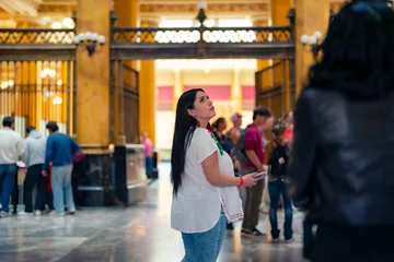 Tuinposter Happy young mexican woman tourist in embroidered top looking around while standing in Postal palace in downtown Mexico City and in light against blurred interior © Itza