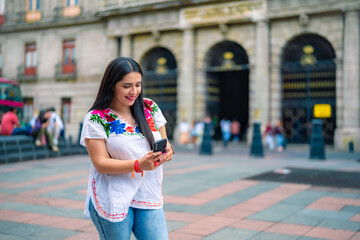 Happy young mexican woman in embroidered top looking at screen of smartphone while standing in...