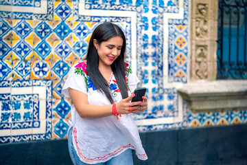 Happy young mexican woman in embroidered top looking at screen of smartphone while standing in front of Sanborns restaurant in downtown Mexico City and reading text messages in daylight