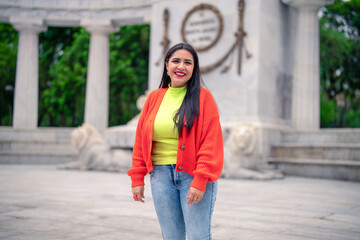 Fototapeta premium Smiling young Hispanic ethnic woman tourist smiling and looking away while standing in street in front of blurred Benito Juarez Hemicycle in Mexico city