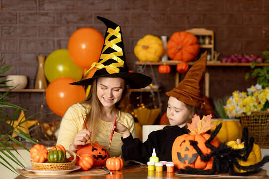 halloween, mom and child daughter in a witch costume with pumpkins and a big spider in a dark kitchen paint a festive pumpkin with paints drawing a muzzle, smile, rejoice