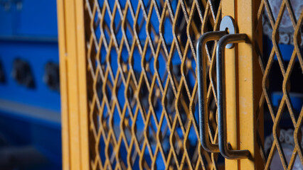 Close-up of a yellow wire mesh door with a silver handle, partially obscuring a blurred blue background.