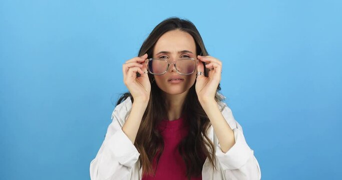 Girl with poor vision looking at camera and rejoicing after putting on eyeglasses isolated against blue background. Young brunette woman with vision problems seeing perfectly with eyeglasses. Try see.