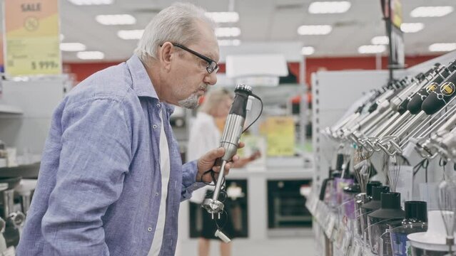 An elderly man in an electronics hypermarket chooses a blender. A pensioner chooses household appliances in a shop