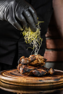The chef decorates a micro-green dish of fried meat on a wooden board on a dark background