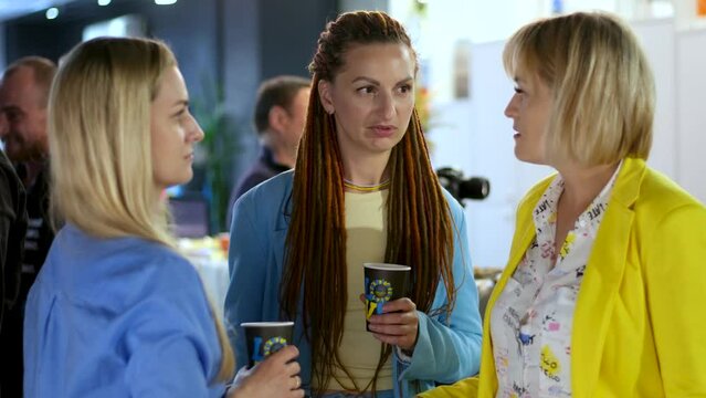 Three women at a corporate party dressed in blue and yellow with a plastic paper cup in their hands communicate, talk, have a nice time, Two blondes and a woman with red dreadlocks