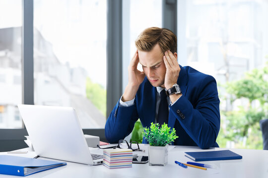 Photo - stressed, tired businessman in suit, heaving headache, hold his head, work with laptop at office. Business fall, job and education concept. Executive employee sitting near window on background