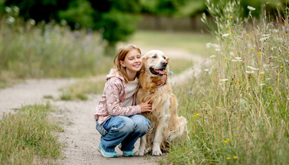 Fototapeta premium Preteen girl with golden retriever dog sitting at nature and looking back. Cute child kid hugging purebred pet doggy in park at summer