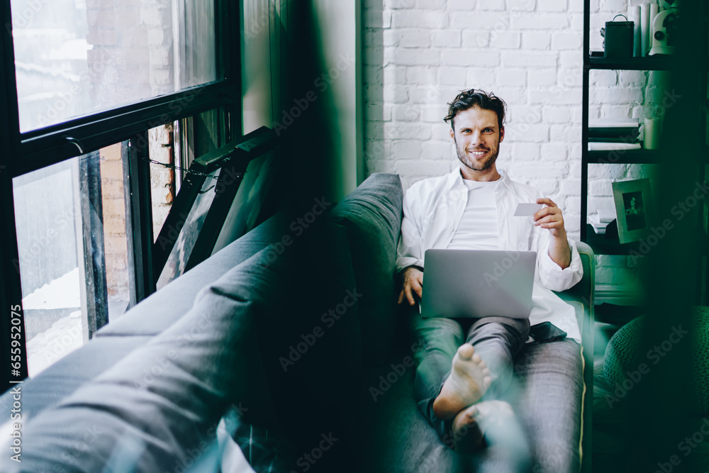 Poster smiling hipster guy buying in web store sitting at sofa in flat