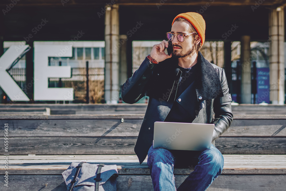 Poster millennial man sitting with netbook and talking via cellphone