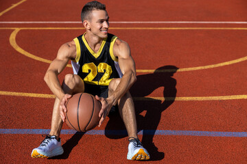 Smiling attractive man sitting on the basketball court after playing game, resting after match.