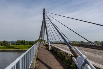 Pedestrian or cycling path parallel to rural road on Lanaye cable-stayed bridge, tower, pylon, huge turnbuckles and cables, trees, misty blue sky in background, wild plants, sunny day in Vise, Belgium