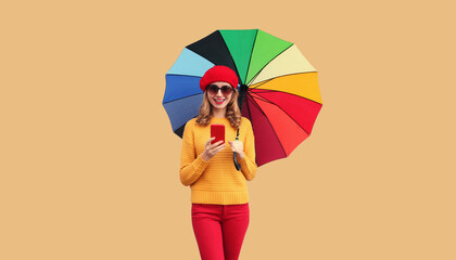 Autumn color style outfit, happy cheerful young woman looking at mobile phone with colorful umbrella wearing red french beret hat, yellow sweater on beige studio background