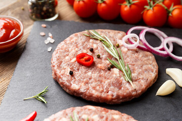 Raw beef hamburger patties on stone plate, vegetables and spices, wooden background.