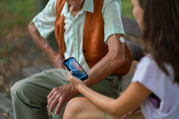 Caregiver helping senior diabetic man check his glucose data on smartphone outdoors, in park.
