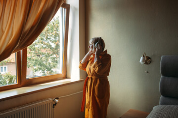An elderly positive woman listens to music in wireless headphones and dances, in the bedroom by the window