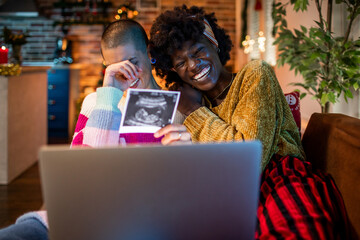 Happy young interracial lesbian couple showing the x ray of their new child on a video call on a laptop in their home