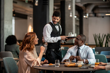 Young waiter serving two diverse business coworkers at the hotel cafe or bar