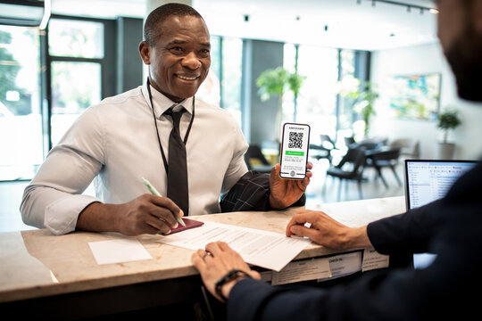 Young African American businessman showing the certificate of his vaccination on a smartphone at the reception of a hotel