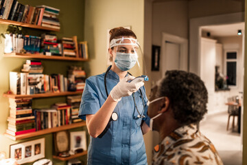 Young female caregiver fever screening her senior patient at their home