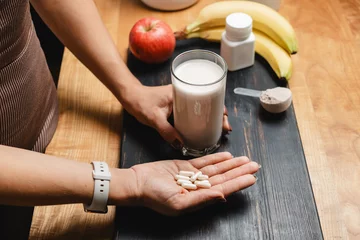 Foto op Canvas Athletic woman in sportswear holding glass of protein drink cocktail and white capsules of amino acids on a table with measuring spoon of protein powder and fruits © O.Farion