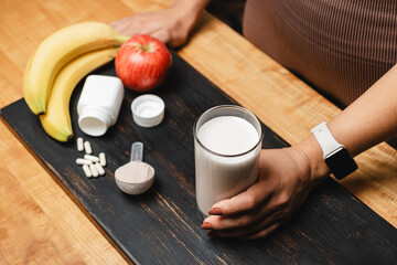 Athletic woman in sportswear holding glass of protein drink cocktail on a table with white capsules of amino acids, measuring spoon of protein powder and fruits
