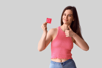 Young woman with condom showing thumb-up on light background