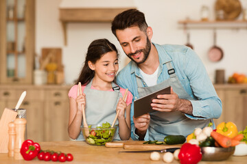 Happy european dad and his daughter cooking in the kitchen, smiling man using digital tablet