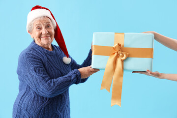 Senior woman in Santa hat receiving Christmas gift on blue background