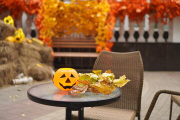 an orange pumpkin basket with a smiling face for sweets on a glass table against the background of an autumn decor with yellow leaves and pumpkins in a cafe on the street, decoration for the Halloween