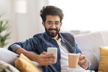 Domestic Leisure. Young Indian Man Using Smartphone And Drinking Coffee At Home