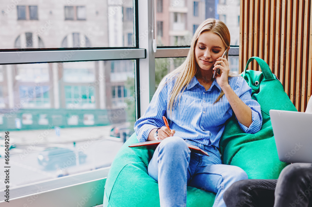 Poster hipster girl calling on smartphone and noting information in notebook