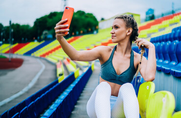 Braided sporty young woman capturing selfie by mobile in grandstand