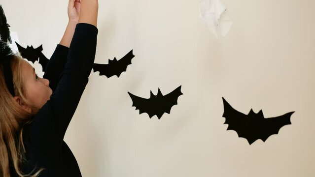 Toddler girl wearing bat costume decorates the house for Halloween party of spooky handmade garland, kid preparing for Halloween and hanging garland on wall. crafts for kids. home decoration concept