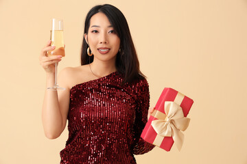 Beautiful Asian woman with champagne and Christmas gift on beige background