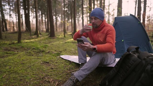 Morning at campground in fall forest. Tursit is browsing Internet on his smartphone while sitting on hiking mat near tent and drinking a beverage from a cup. Travel, people and healthy lifestyle. 