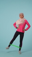 Full-sized isolated photo capturing an attractive young woman wearing a sportswear and a hijab, sheila. She is exercising with a green rubber exercise band.