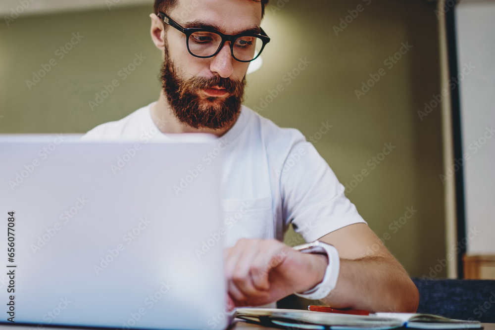 Poster hipster guy in spectacles checking time planning schedule using netbook for studying