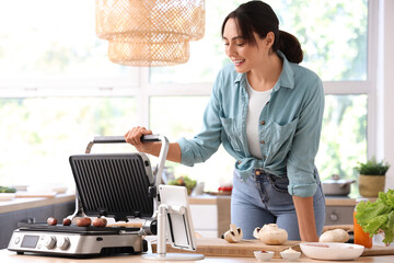Beautiful young woman cooking delicious sausages on modern electric grill in kitchen