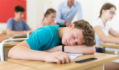 Tired teen student sleeping at desk in classroom during lesson on blurred background of classmates ..