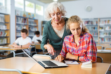 Mature female teacher working with schoolchildren in the library cheks the assignment from a European fifteen-old-year schoolgirl