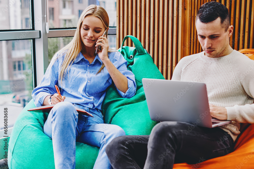 Poster hipster girl calling on smartphone and noting information in notebook sitting on pouf while fer boyf