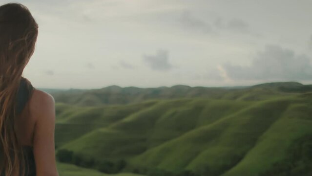 Woman back view look at green hills meadow nature landscape. Dramatic evening shot tourist woman stand with long hair blowing in wind. Outdoor lifestyle travel on summer holiday vacation. Slow motion