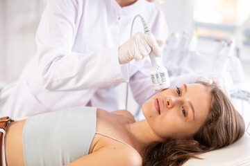 Positive young girl undergoing facial treatment using cold hammer helping to shrink pores and...
