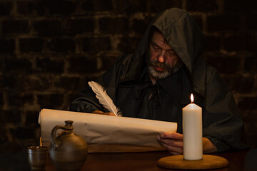 A monk with a beard writes with a quill pen on parchment against a dark brick wall, dark tonality.