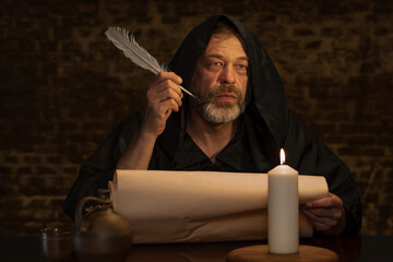 An old monk with a beard holds a quill in his hand over a parchment against a dark brick wall, dark...